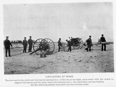 LIFE-SAVERS AT WORK
The two men in the center are burying the sand-anchor; of the two at the right, one is ready with the crotch support the hawser and the other carries the breeches-buoy; the other three men are hauling the line which has already been shot over the wrecked vessel.